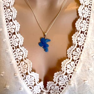 Puede incluir: Un collar de plata con un colgante azul con forma de p&aacute;jaro estilizado. El colgante cuelga de una delicada cadena. El collar se muestra sobre una blusa blanca con ribete de encaje.