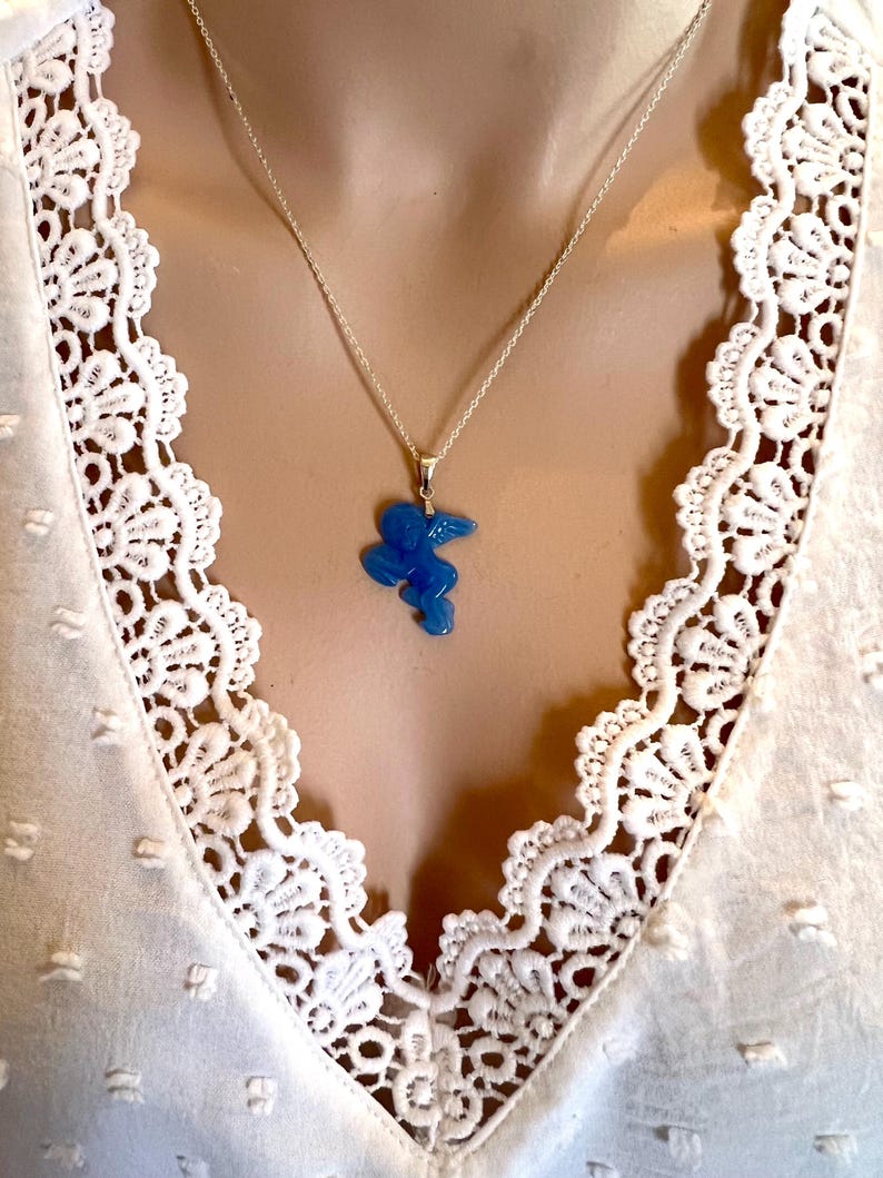 Puede incluir: Un collar de plata con un colgante azul con forma de p&aacute;jaro estilizado. El colgante cuelga de una delicada cadena. El collar se muestra sobre una blusa blanca con ribete de encaje.