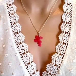 Puede incluir: Un collar de plata con un colgante rosa brillante en forma de flor. El collar se lleva sobre una blusa blanca con un delicado ribete de encaje.