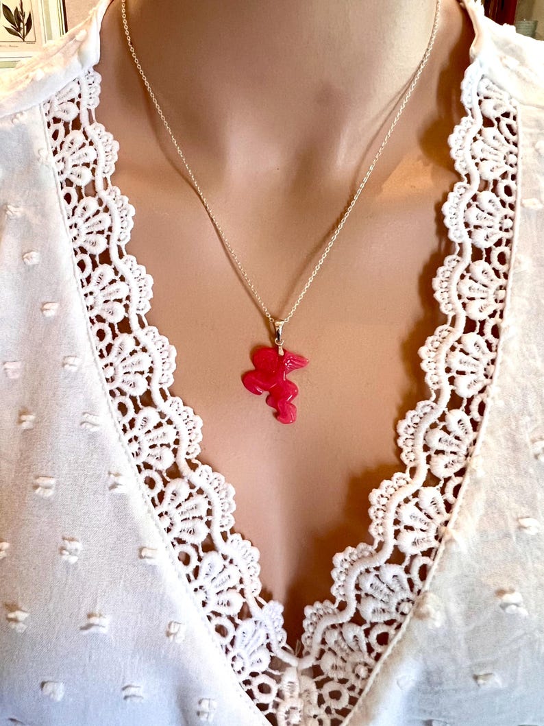 Puede incluir: Un collar de plata con un colgante rosa brillante en forma de flor. El collar se lleva sobre una blusa blanca con un delicado ribete de encaje.