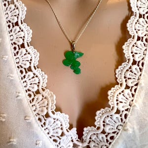 Puede incluir: Un collar de plata con un colgante verde brillante con un dise&ntilde;o floral estilizado. El colgante cuelga de una delicada cadena. El collar se muestra sobre un top blanco con ribete de encaje.