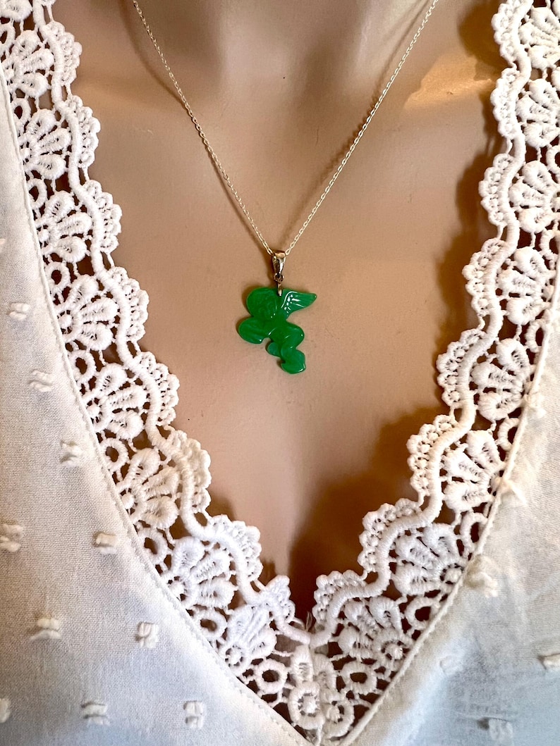Puede incluir: Un collar de plata con un colgante verde brillante con un dise&ntilde;o floral estilizado. El colgante cuelga de una delicada cadena. El collar se muestra sobre un top blanco con ribete de encaje.