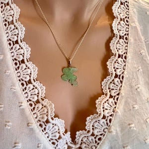 Puede incluir: Un delicado collar de plata con un colgante de jade verde claro con forma de animal estilizado. El colgante cuelga de una fina cadena de plata. El collar se muestra sobre una blusa blanca con ribete de encaje.