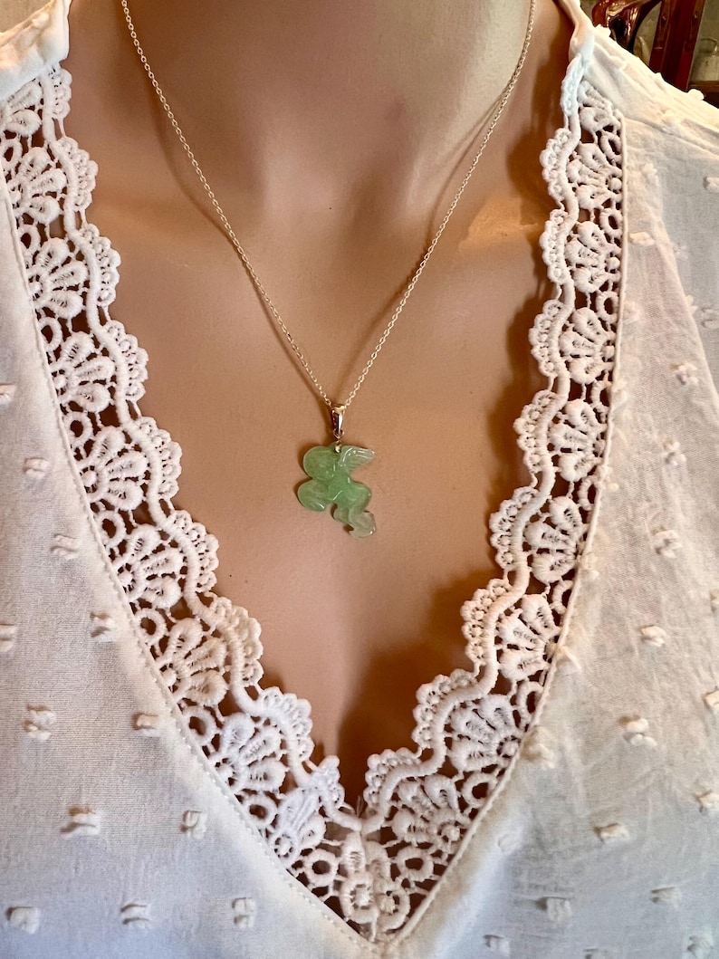 Puede incluir: Un delicado collar de plata con un colgante de jade verde claro con forma de animal estilizado. El colgante cuelga de una fina cadena de plata. El collar se muestra sobre una blusa blanca con ribete de encaje.