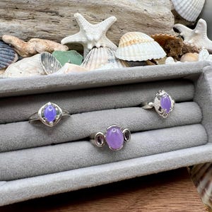 May include: Three silver rings with oval and teardrop-shaped lavender gemstones are displayed in a gray jewelry box. The rings have intricate silver detailing. The background includes seashells, driftwood, and a starfish.