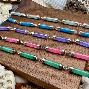 May include: A collection of colorful gemstone bracelets. Each bracelet features a series of cylindrical, polished gemstones in shades of green, blue, purple, pink, and silver connectors. The bracelets are displayed on a wooden surface.