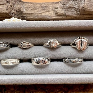 May include: A collection of silver rings displayed in a gray velvet jewelry box. The rings feature various designs, including foot prints, nautical themes, and star patterns. The rings are presented in a wooden box.