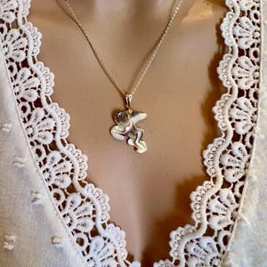 Puede incluir: Un collar con colgante de plata con un dise&ntilde;o abstracto. El colgante cuelga de una fina cadena de plata. El collar se muestra sobre una blusa blanca con detalles de encaje.