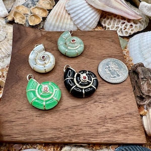 May include: Four seashell-shaped pendants in white, green, and black with red accents, displayed on a wooden surface. A US quarter coin is next to the pendants. The pendants are surrounded by seashells.