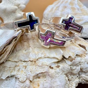 May include: Three silver rings with cross-shaped designs. One ring features a dark blue inlay, while the other two have purple inlays. The rings are displayed on a textured, off-white surface, creating a contrast with the shiny silver.