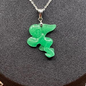 Puede incluir: Collar con colgante verde esmeralda que representa un &aacute;ngel tallado con alas extendidas. La cadena es de plata. El texto "VERDANT GREEN" se muestra en la parte inferior de la imagen.
