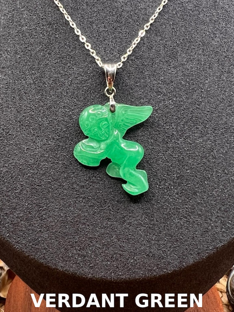 Puede incluir: Collar con colgante verde esmeralda que representa un &aacute;ngel tallado con alas extendidas. La cadena es de plata. El texto "VERDANT GREEN" se muestra en la parte inferior de la imagen.