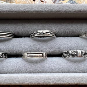 May include: A collection of silver rings displayed in a gray velvet jewelry box. The rings feature various designs, including braided, textured, and banded styles. The rings are made of a shiny, metallic material.