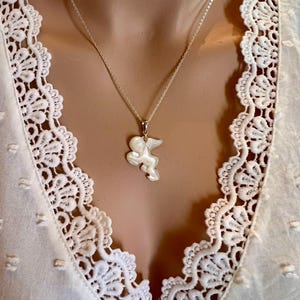 Puede incluir: Un collar de plata con un colgante en forma de querub&iacute;n alado. El colgante blanco cuelga de una fina cadena de plata. El collar se muestra sobre una blusa blanca con detalles de encaje.