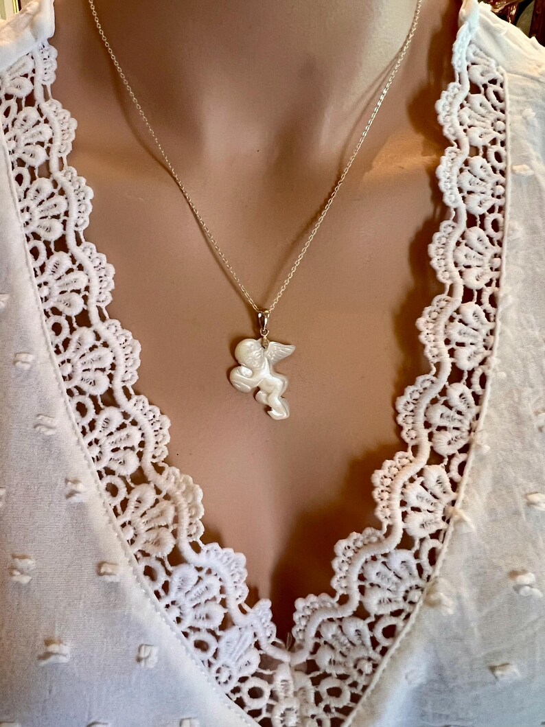 Puede incluir: Un collar de plata con un colgante en forma de querub&iacute;n alado. El colgante blanco cuelga de una fina cadena de plata. El collar se muestra sobre una blusa blanca con detalles de encaje.