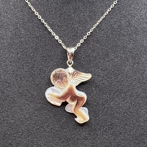 Puede incluir: Un collar de plata con un colgante con forma de querub&iacute;n. El colgante es una mezcla de marr&oacute;n, beige y blanco. El collar se muestra sobre un fondo negro. El texto "GOLDEN TIDE" es visible en la parte inferior de la imagen.