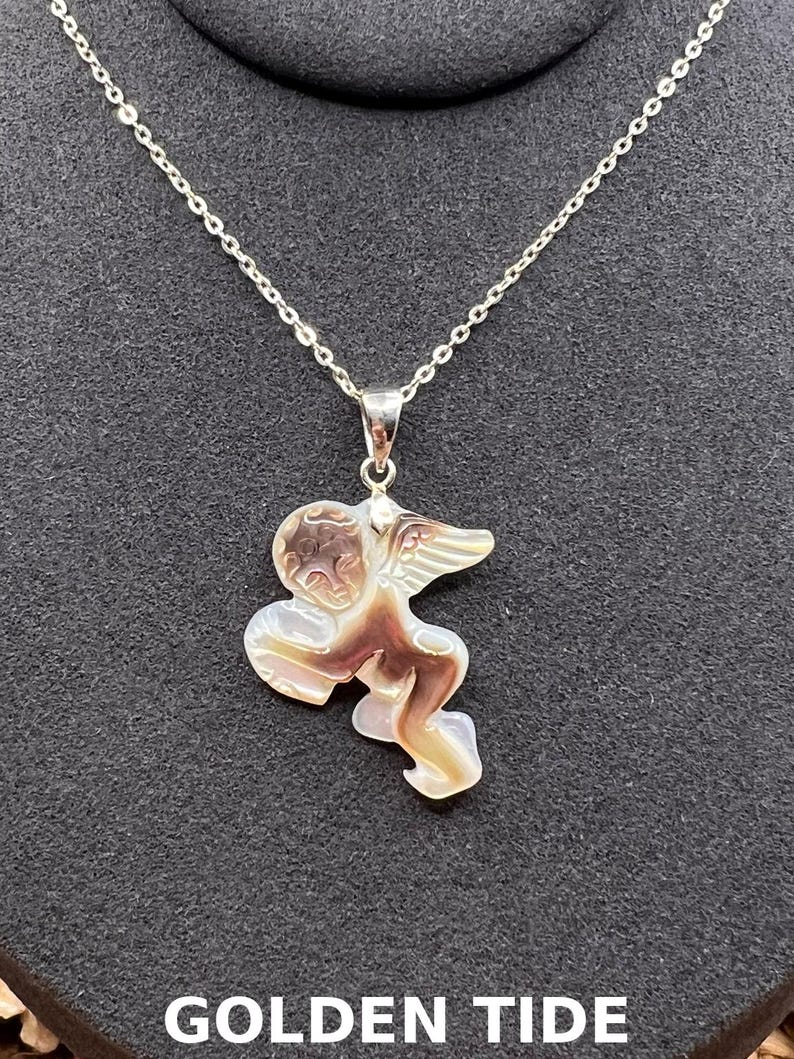 Puede incluir: Un collar de plata con un colgante con forma de querub&iacute;n. El colgante es una mezcla de marr&oacute;n, beige y blanco. El collar se muestra sobre un fondo negro. El texto "GOLDEN TIDE" es visible en la parte inferior de la imagen.