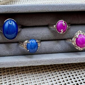 May include: A collection of four rings displayed in a gray jewelry box. The rings feature oval and teardrop-shaped gemstones in vibrant blue and magenta hues, set in silver bands. The gemstones have a polished, glossy finish.