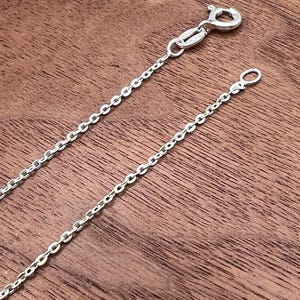 Puede incluir: Un delicado collar de cadena de plata con un cierre de mosquet&oacute;n y un peque&ntilde;o lazo en el extremo. La cadena est&aacute; compuesta por peque&ntilde;os eslabones ovalados espaciados. El collar se muestra sobre una superficie de madera.