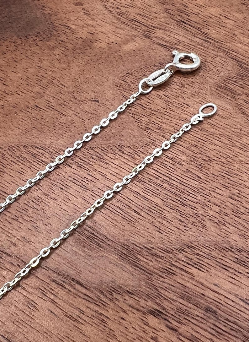 Puede incluir: Un delicado collar de cadena de plata con un cierre de mosquet&oacute;n y un peque&ntilde;o lazo en el extremo. La cadena est&aacute; compuesta por peque&ntilde;os eslabones ovalados espaciados. El collar se muestra sobre una superficie de madera.