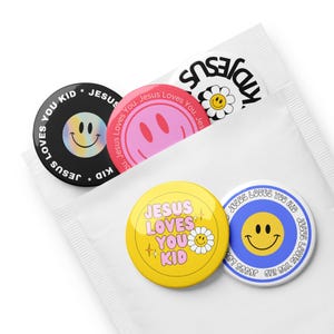 May include: A collection of round buttons with smiley faces and the text "JESUS LOVES YOU KID". The buttons are in various colors, including black, pink, yellow, and blue, with white text and a daisy graphic.
