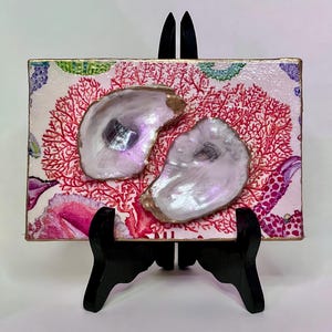 May include: Decorative rectangular art piece featuring two iridescent oyster shells with gold edges. The shells are set against a pink and red coral-like background with floral accents. The artwork is displayed on a black easel.
