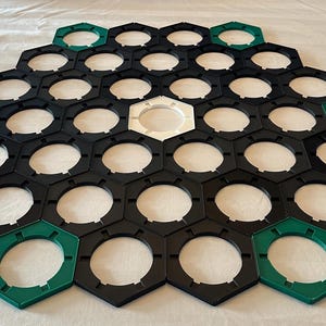 Puede incluir: Un tablero de juego hexagonal compuesto por piezas negras, blancas y verdes. El tablero está formado por baldosas entrelazadas en forma de panal. Las baldosas verdes se colocan alrededor del perímetro. La baldosa central es blanca.