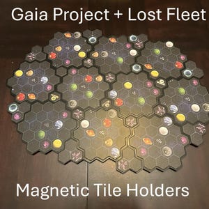 Op de afbeelding: Een verzameling zeshoekige magnetische tegelhouders voor de bordspellen Gaia Project en Lost Fleet. De tegels zijn zwart met kleurrijke planeet- en sterrenafbeeldingen. De tekst "Gaia Project + Lost Fleet" en "Magnetic Tile Holders" is zichtbaar.