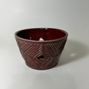 May include: A small, red ceramic planter with a geometric pattern and a drainage hole in the bottom.