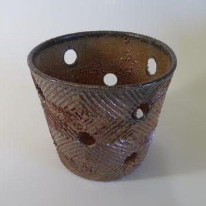 May include: A handcrafted ceramic pot with a textured surface and a series of circular openings. The pot features a brown and reddish-brown color scheme with a dark rim. The design includes diagonal etched lines.