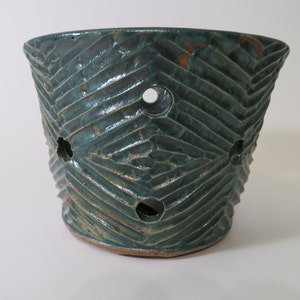 May include: A handcrafted teal ceramic planter with a textured, chevron-like pattern. The planter features several circular drainage holes and a rustic, earthy aesthetic. The planter is suitable for indoor or outdoor use.