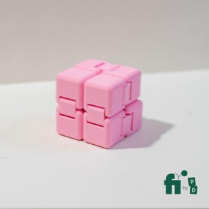 May include: A pink infinity cube fidget toy. The cube is made of several connected smaller cubes that can be flipped and folded. The toy is a solid, pastel pink color and sits on a white surface.