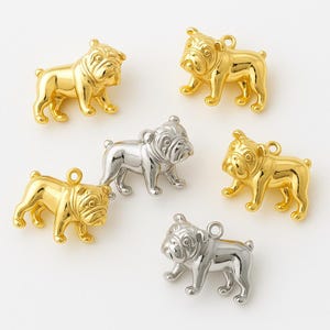 Puede incluir: Una colección de dijes de bulldog dorados y plateados. Los dijes son detallados, con arrugas visibles y un lazo para sujetarlos a la joyería. Los bulldogs están de pie, mostrando sus rasgos distintivos.