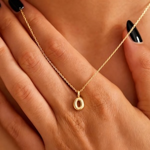 Puede incluir: Un collar dorado con un colgante con forma de la letra "O". El colgante es pequeño y redondo, con un acabado pulido. El collar es delicado y parece estar hecho de una cadena fina. El fondo es la piel de una persona.