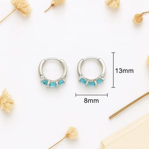 Puede incluir: Un par de pendientes de aro plateados, cada uno adornado con tres gemas de color azul claro. Los pendientes miden 8 mm de ancho y 13 mm de diámetro. La imagen está sobre un fondo blanco con flores secas.