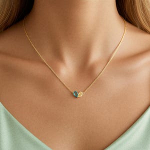 Puede incluir: Delicado collar dorado con una fina cadena, con dos gemas en forma de lágrima, una azul y otra amarilla. El colgante cuelga de una fina cadena dorada. El fondo es de color verde claro.