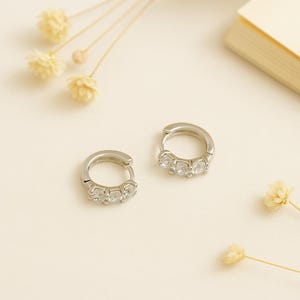 Puede incluir: Dos pendientes de aro plateados, cada uno adornado con tres gemas redondas y transparentes. Los pendientes se exhiben sobre una superficie de color crema, con flores secas y una pequeña pila de papel en el fondo.