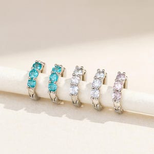 Puede incluir: Cinco pendientes de aro plateados, cada uno adornado con cuatro piedras preciosas. Las piedras son de color turquesa, transparente y rosa. Los pendientes se muestran sobre una superficie blanca, sobre un fondo neutro.