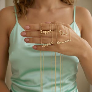 Puede incluir: Una mano muestra varios collares dorados con nombres. Los nombres "Copper", "Jessica", "Olivia" y "Alexandra" son visibles. Los collares tienen cadenas delicadas y se exhiben sobre un fondo verde claro.