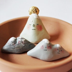 May include: Three whimsical ceramic mountain figurines with painted faces and snow-capped peaks. One mountain has a gold-colored top. The mountains are in a terracotta-colored bowl.