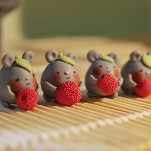 May include: Four gray mouse figurines with red berry-like objects. Each mouse has a green leaf on its head and rosy cheeks. The figurines are arranged in a row on a light brown surface, creating a whimsical and charming scene.