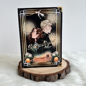 Puede incluir: Un libro de tapa dura titulado "Greenlight" con una cubierta negra que presenta dos figuras ilustradas y detalles dorados. La portada incluye fuegos artificiales, flores de naranjo y el texto "Greenlight". El libro se exhibe sobre una rodaja de madera.