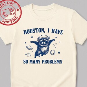 May include: Cream-colored t-shirt featuring a navy blue illustration of a raccoon in an astronaut helmet, with the text "Houston, I Have So Many Problems." The design includes stars, planets, and a sun. The shirt has a round neckline and short sleeves.