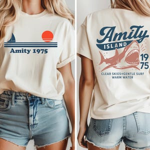 May include: Cream-colored t-shirt with a nautical design on the front, featuring a sailboat, a red sun, and the text "Amity 1975." The back of the shirt displays a shark graphic with the words "Amity Island 1975" and "Clear Skies+Gentle Surf Warm Water."