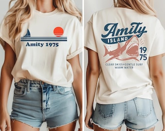 Puede incluir: Camiseta color crema con un diseño náutico en la parte delantera, con un velero, un sol rojo y el texto "Amity 1975". La parte trasera de la camiseta muestra un gráfico de tiburón con las palabras "Amity Island 1975" y "Clear Skies+Gentle Surf Warm Water".