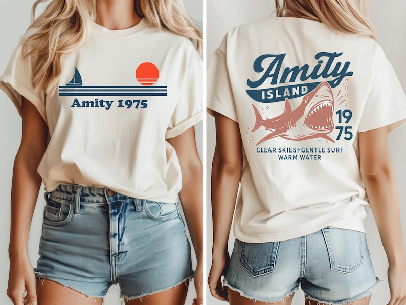 May include: Cream-colored t-shirt with a nautical design on the front, featuring a sailboat, a red sun, and the text "Amity 1975." The back of the shirt displays a shark graphic with the words "Amity Island 1975" and "Clear Skies+Gentle Surf Warm Water."