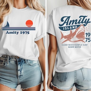 May include: White t-shirt with a nautical design featuring a sailboat, sun, and the text "Amity 1975" on the front. The back of the shirt displays a shark graphic with the words "Amity Island 1975" and "Clear Skies+Gentle Surf Warm Water."