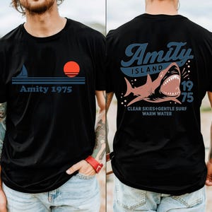 May include: Black t-shirt with a nautical design featuring a sailboat, sun, and the text "Amity 1975" on the front. The back of the shirt displays a shark graphic with the words "Amity Island 1975" and "Clear Skies + Gentle Surf Warm Water."