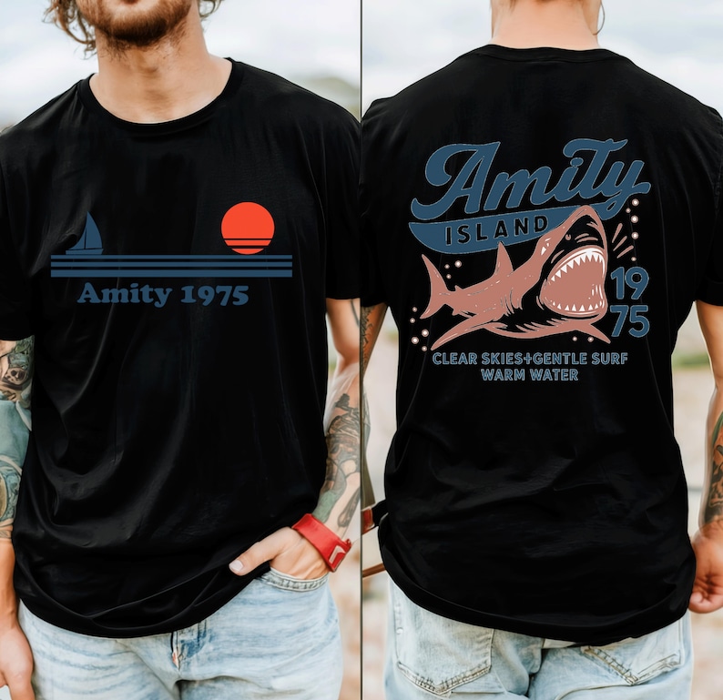 May include: Black t-shirt with a nautical design featuring a sailboat, sun, and the text "Amity 1975" on the front. The back of the shirt displays a shark graphic with the words "Amity Island 1975" and "Clear Skies + Gentle Surf Warm Water."