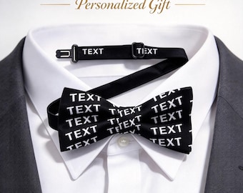 May include: A black bowtie featuring the word TEXT printed in white repeatedly across the fabric. The bowtie includes an adjustable black neck strap with a metal hook and slider. It is displayed over a white collared shirt and a dark gray blazer.